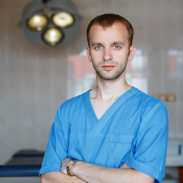 Young attractive male doctor in blue medical clothing is standing in the hospital in the operating room against the background of medical equipment. Modern medicine and patient care. - Zdjęcie, obraz