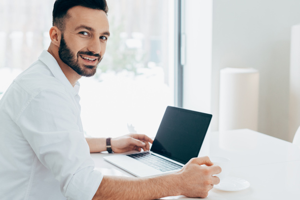 Smiling man in white shirt looking at camera while using laptop with blank screen - Foto, Bild