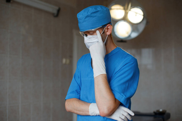 Young professional doctor in blue uniform with glasses with gloves in a mask is thinking in the operating room on the background of a bright lamp. Surgeon is preparing for surgery. Modern medicine. - Fotoğraf, Görsel