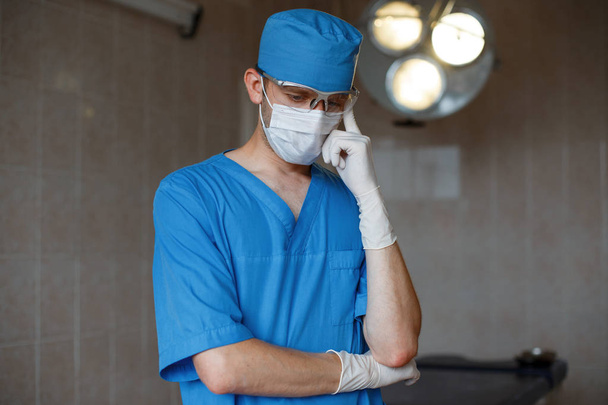 Il giovane chirurgo in uniforme medica blu con una maschera bianca protettiva che indossa guanti di gomma è in piedi in ospedale e pensa. Il neurochirurgo si sta preparando per l'intervento. Nozione di assistenza sanitaria
. - Foto, immagini