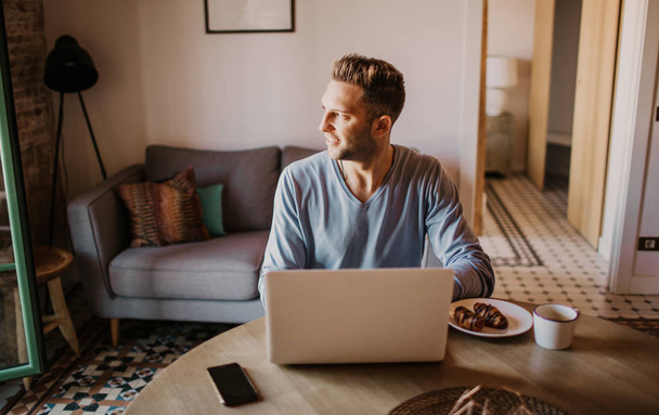Handsome coworker man working at living room at home. Man sitting at wooden table using laptop during breakfast at kitchen. Blurred background. - 写真・画像