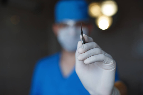 Der plastische Chirurg in blauer Uniform mit medizinischer Maske und Skalpell in der Hand beginnt zu handeln. Facharzt im Operationssaal. - Foto, Bild