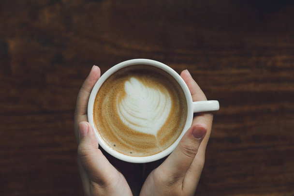 Женщина держит за руку кружку белого кофе. Кофе это латте. стол на деревянном столе в винтажном стиле, взятом с верхнего вида, смотрите на молочную пену
. - Фото, изображение