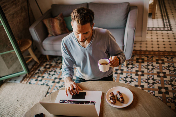 Handsome coworker man working at living room at home. Man sitting at wooden table using laptop during breakfast at kitchen. Blurred background. - Foto, imagen