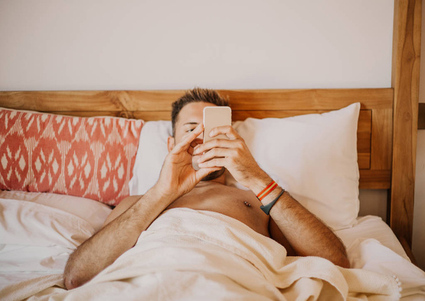 Carefree gay enjoying new day.Handsome young man in bed typing on cell phone, sending text message or dialing number - 写真・画像