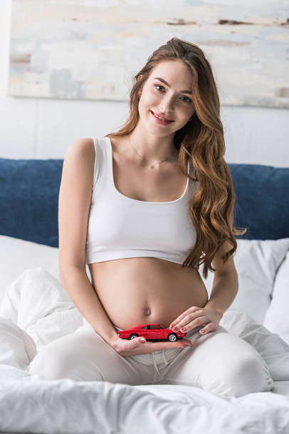 Blissful pregnant woman sitting on bed and holding red toy car - Fotografie, Obrázek