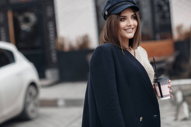 A young beautiful woman with long,hair and gray eyes,beautiful makeup,pink plump lips,sweet smile,brunette,spending her time outdoors in the spring in the city,wearing a white knitted sweater,posing in the town.Portrait of happy young woman in a cap - Fotografie, Obrázek