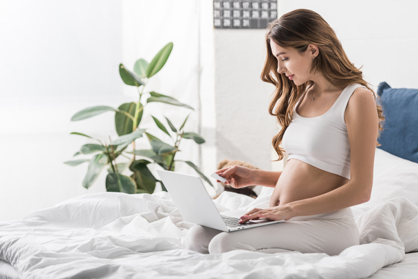 Güzel hamile kadın yatakta laptop ve kredi kartı ile oturuyordu - Fotoğraf, Görsel