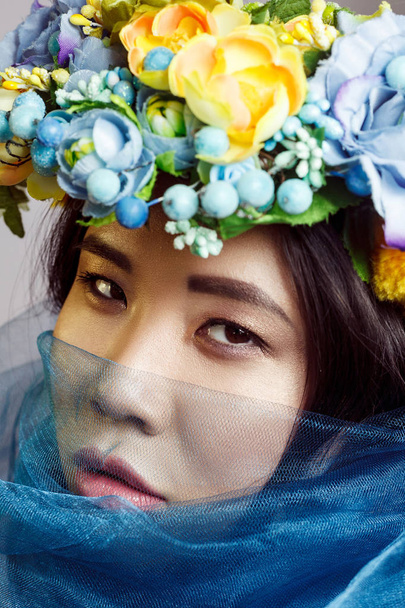 portrait of asian woman with floral hat and blue veil looking at camera on light grey background - Foto, afbeelding