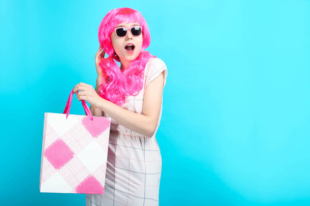 Hermosa chica sorprendida con pelo rosa brillante en vestido con bolsa de compras sobre fondo azul. Concepto venta, compras
. - Foto, Imagen