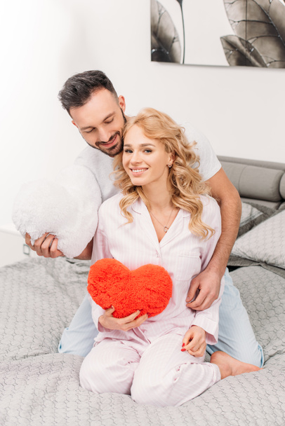 Happy couple sitting on bed and holding toy hearts - 写真・画像
