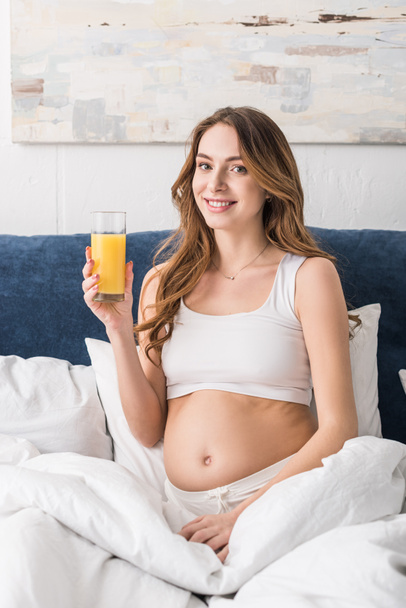 Vidám terhes nő ül ágyas és a gazdaság pohár gyümölcslé - Fotó, kép