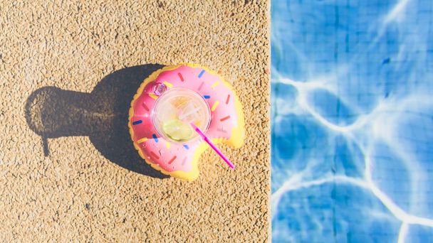 Tasty refreshing cocktail on on inflatable donut plastic toy on edge of swimming pool. Ripple Water in swimming pool with sunny reflection.Wide. Top view. - Photo, image