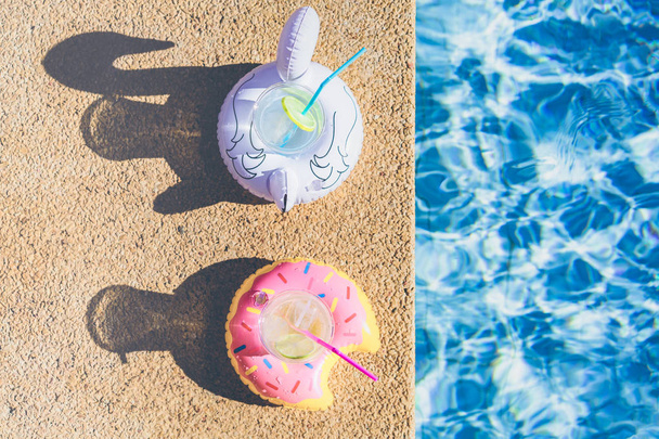 Twee heerlijke verfrissende cocktail op opblaasbare donut y witte flamingo plastic speelgoed op rand van zwembad. Water in zwembad met zonnige reflectie rimpel. Bovenaanzicht. - Foto, afbeelding