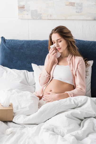 Upset pregnant woman sitting on bed and wiping tears with napkin - Фото, изображение