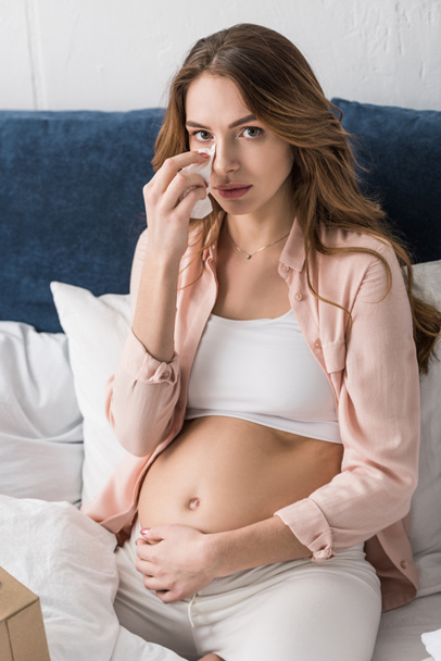 Грустная беременная девушка вытирает лицо салфеткой, сидя на кровати
 - Фото, изображение