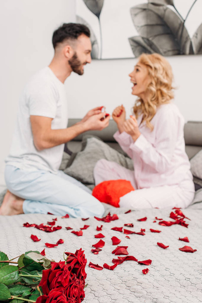 Man with beard sitting on bed with rose petals and making proposal to girlfriend - Foto, afbeelding
