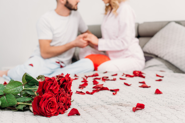 Partial view of couple sitting on bed with rose petals and holding hands - Фото, изображение