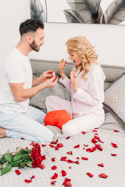 Man sitting on bed with rose petals and proposing to curly girl - Фото, зображення