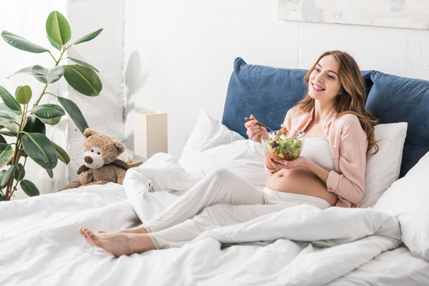 Вдохновлённая беременная женщина ест салат в постели
 - Фото, изображение