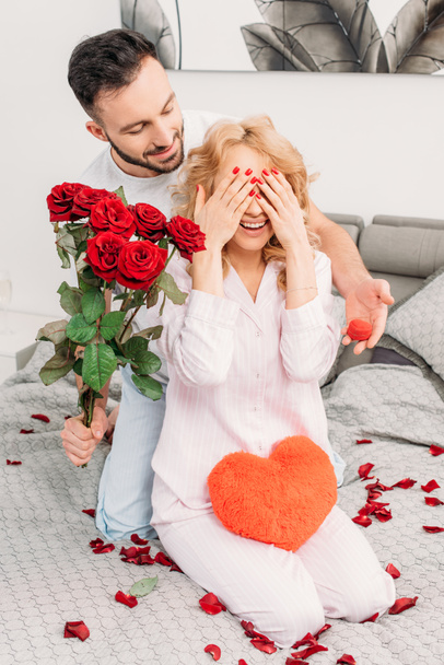 Man making proposal and presenting flowers to girlfriend in bedroom - Фото, изображение