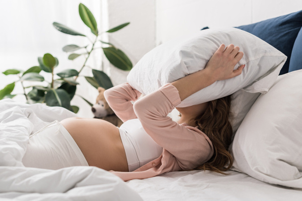 Mujer embarazada acostada en la cama y con almohada en la cabeza
 - Foto, Imagen