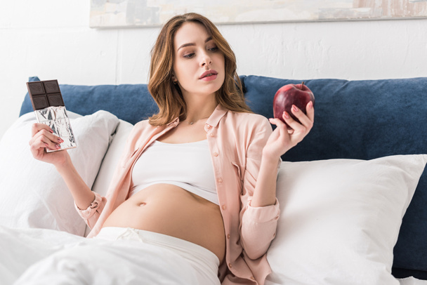 Mujer embarazada sosteniendo manzana roja y barra de chocolate
 - Foto, Imagen