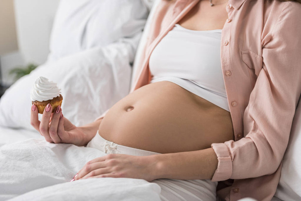 Partial view of pregnant woman with cupcake lying in bed - Photo, image