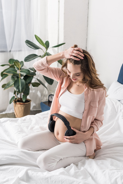 Pregnant woman sitting on bed and holding headphones on belly - Foto, afbeelding