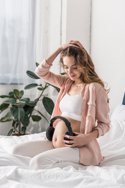 Jolie femme enceinte assise sur le lit avec un casque sur le ventre
 - Photo, image