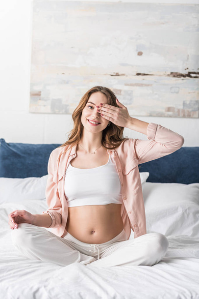 Blissful pregnant woman sitting on bed and covering eye with hand - Foto, Imagem