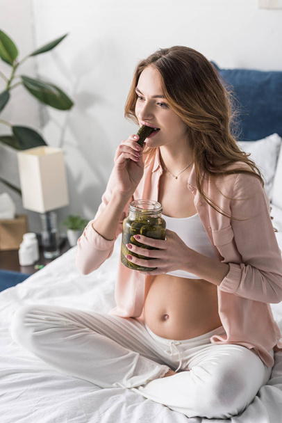 Brunette zwangere vrouw eten augurken in bed - Foto, afbeelding