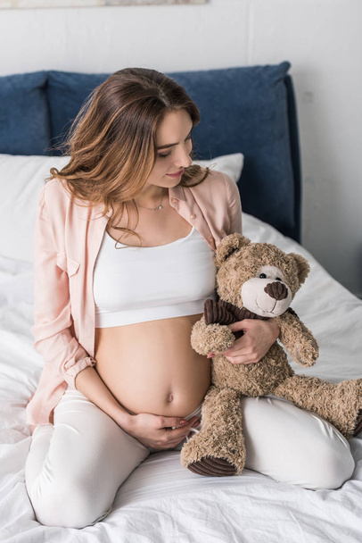 Беременная молодая женщина сидит на кровати и смотрит на игрушечного медведя
 - Фото, изображение