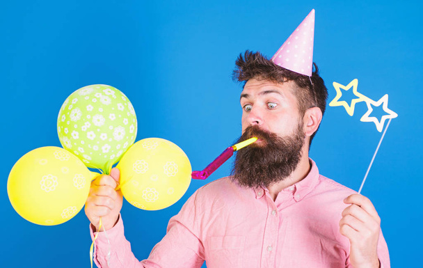 Hipster with star shaped glasses blows into party horn. Surprise concept. Guy in party hat with holiday attributes celebrates. Man with beard on surprised face holds air balloons, blue background - Fotoğraf, Görsel