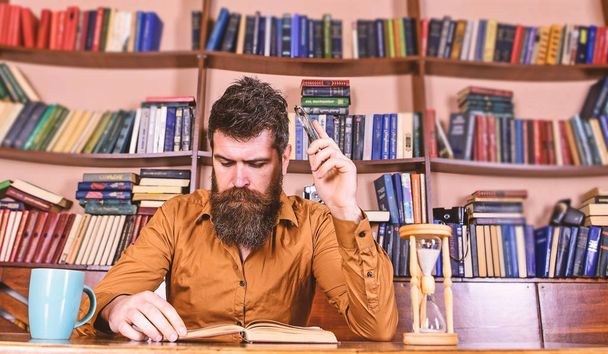 Man on busy face reading book, bookshelves on background. Education and science concept. Teacher or student with beard studying in library. Scientist reading, sits at table with mug and hourglass - Foto, Imagen