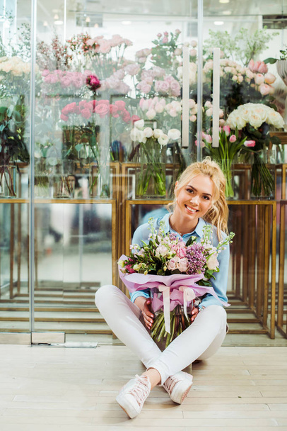 bela mulher sorridente segurando buquê de flores enquanto sentado na frente da loja de flores com espaço de cópia
 - Foto, Imagem