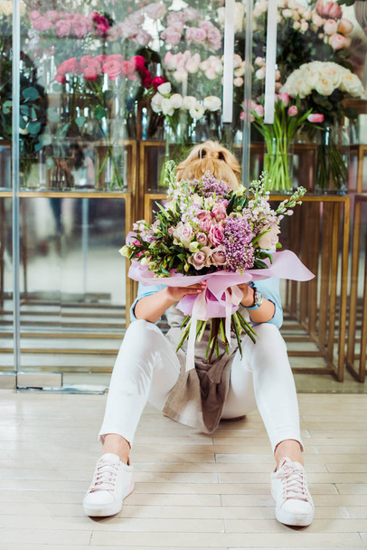 γυναίκα κρατώντας την ανθοδέσμη λουλουδιών μπροστά από το πρόσωπό ενώ κάθεστε δίπλα στο ανθοπωλείο  - Φωτογραφία, εικόνα