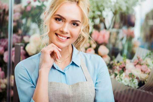 beautiful female florist in apron touching ching and smiling with flower shop on background - Foto, Imagem