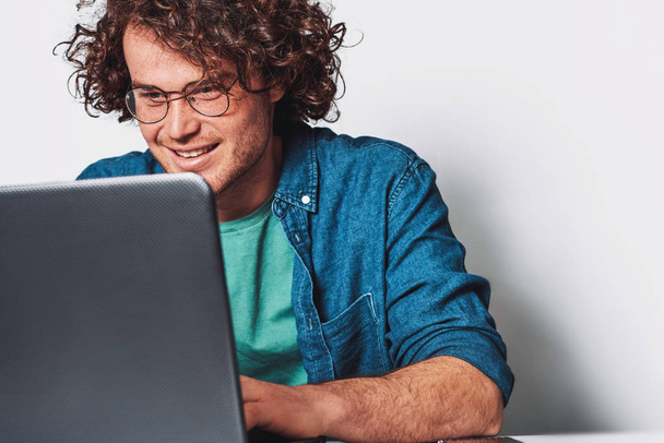 Young happy businessman with curly hair working on his laptop in office. Smiling freelancer man sitting at his desk surfing internet on laptop computer. - Фото, изображение