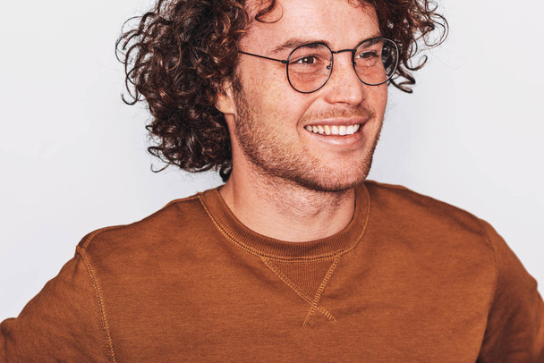 Closeup horizontal portrait of handsome smiling young male with curly hair, wears brown pullover and round trendy spectacles, looking to the camera. Business man posing on white studio wall. Copyspace - Valokuva, kuva