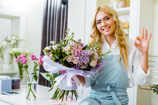 красивая улыбчивая хозяйка цветочного магазина, сидя на прилавке и держа в руках букет, показывая хороший знак
 - Фото, изображение