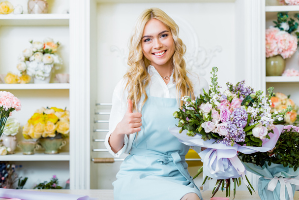 beautiful smiling female flower shop owner sittng on counter and holding bouquet while showing thumb up sign - Φωτογραφία, εικόνα