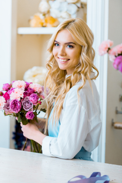 beautiful female florist holding bouquet with roses and carnations in flower shop and looking away - Фото, зображення