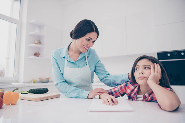 Portrait of two nice cute winsome lovely attractive cheerful mom helping annoyed bored tired girl doing courses classes in light white kitchen interior indoors - Фото, зображення