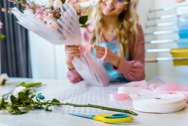 partial view of female florist arranging bouquet in flower shop with ribbon and scissors on foreground - Фото, изображение