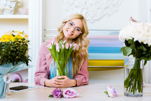 красивая флористка смотрит в камеру и держит вазу с розовыми тюльпанами в цветочном магазине
 - Фото, изображение
