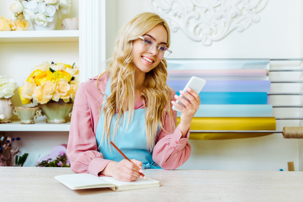 belle femme souriante propriétaire de magasin de fleurs dans des lunettes écrit dans un cahier et en utilisant smartphone
 - Photo, image