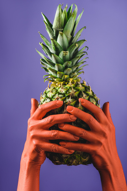 bijgesneden beeld van vrouw met rijpe ananas fruit in koraal gekleurde handen op een violette achtergrond, kleur van 2019 concept - Foto, afbeelding