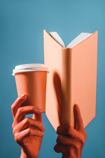 cropped view of coral colored female hands with coral paper cup and book on blue background, color of 2019 concept - Photo, Image