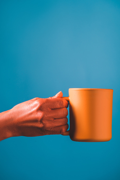 partial view of coral colored female hand and cup on blue background, color of 2019 concept - Photo, Image